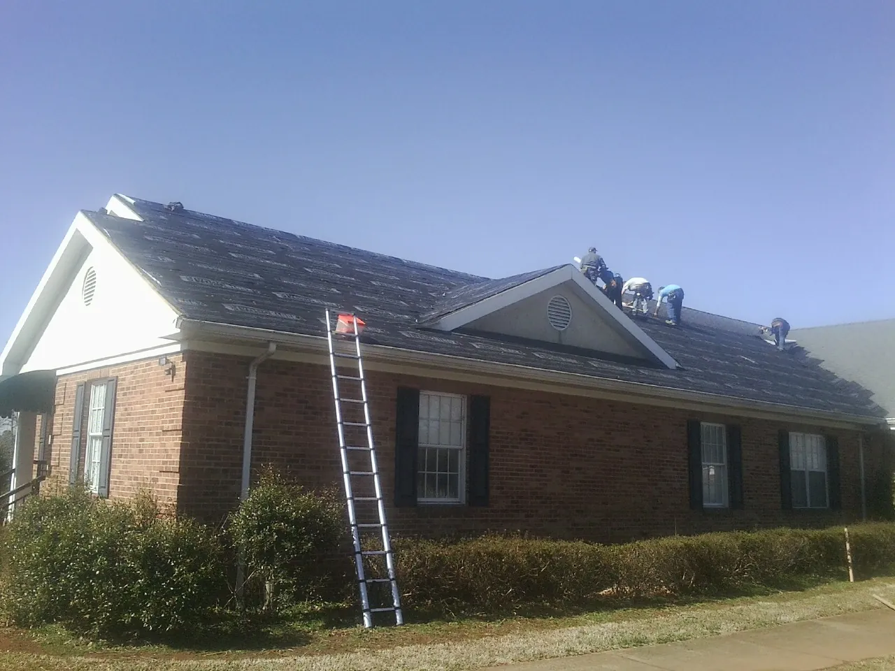 Crew tearing shingles off a brick ranch roof with a ladder set — Emergency Roof Repair work by our crew in Tribune