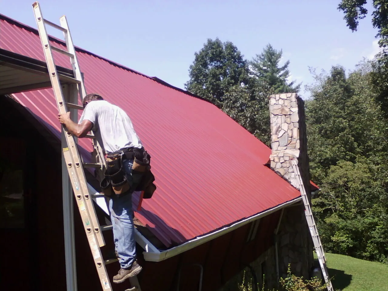 Roofer on a ladder beside a red metal roof and stone chimney — Soffit & Fascia Repair by Roofing Brothers in Tribune