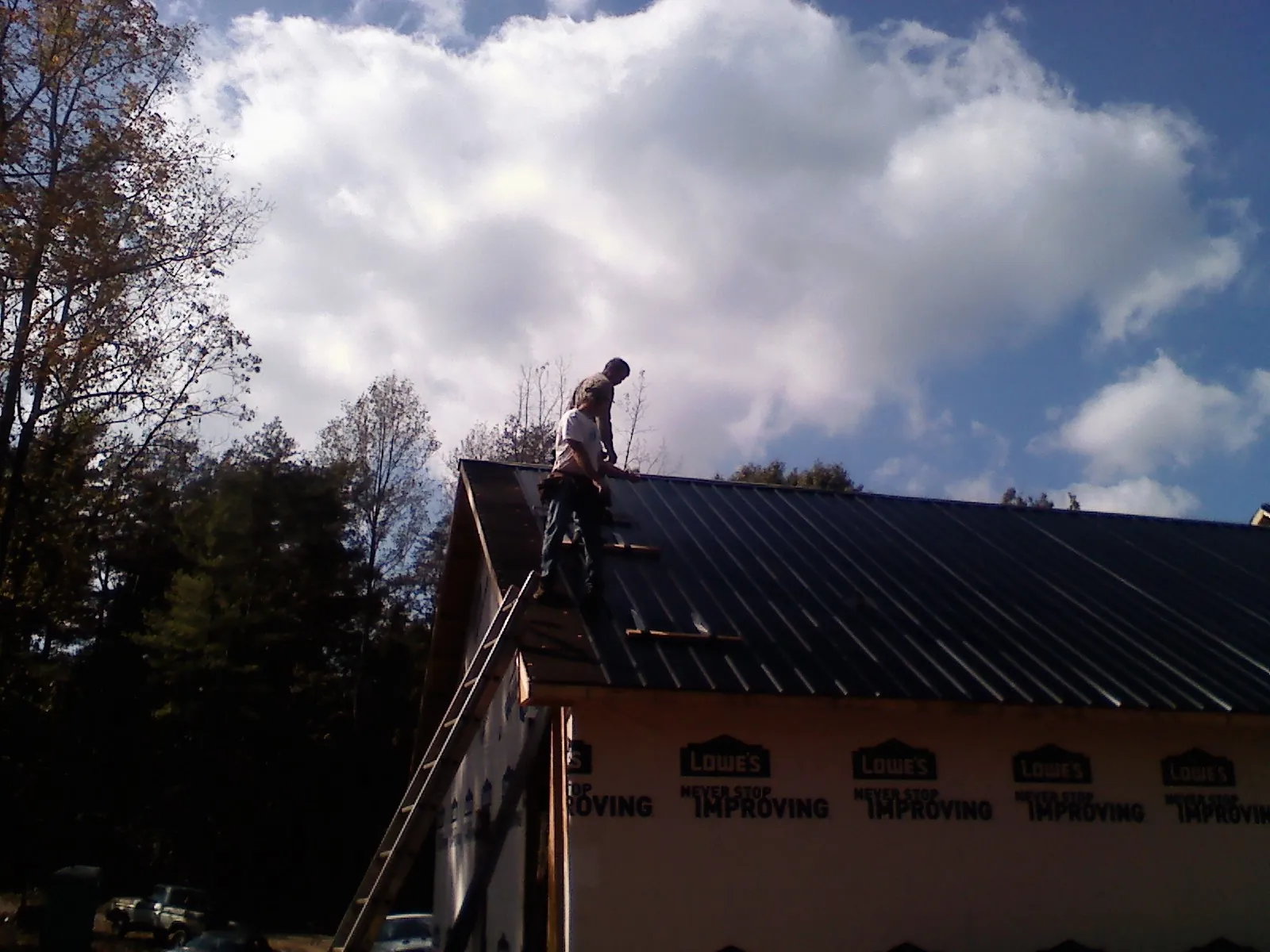 Roofer fastening dark metal panels high against a cloudy sky, from a Roof Inspection job in Tribune